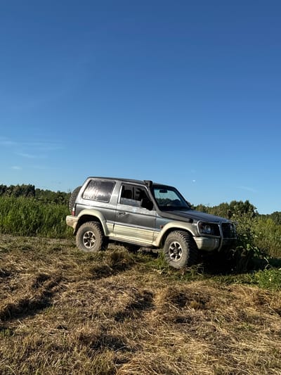 An off-road vehicle, a grey and beige Mitsubishi Pajero, is parked on a field of dry, mowed grass under a clear blue sky. The vehicle is angled towards the right of the frame, with its spare tire mounted on the rear door. Tall green grass and trees form a backdrop to the right of the vehicle, while a line of trees is visible in the distance to the left. The ground in the foreground is uneven and covered with a layer of dry, cut vegetation, suggesting a rural or outdoor setting, possibly in or near the village of Tatishchev Pogost, Russia. There are no people or animals visible in the image.