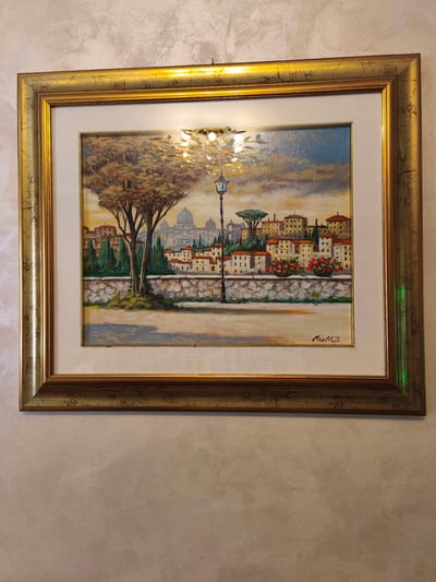 The image shows a framed painting of a cityscape, likely Rome, Italy, due to the prominent dome in the background resembling St. Peter's Basilica. The painting depicts a street scene with buildings, trees, a lamppost, and a stone wall. The style appears to be oil on canvas. The painting is framed in an ornate gold-colored frame and hangs on a textured, neutral-colored wall. There is no information in the image to suggest the location is Campochiaro, Italy.