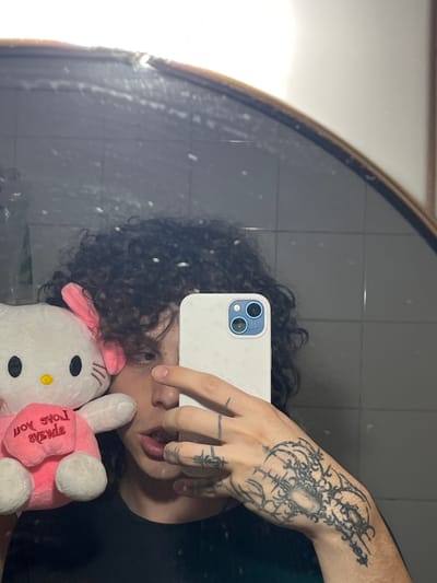 A person with curly hair is taking a selfie in a mirror. The person is holding a white iPhone with multiple cameras and is sticking out their tongue. Their hand has a large, intricate tattoo on the back and smaller tattoos on the fingers. They are holding a Hello Kitty plush toy. The background shows grey wall tiles. It is not possible to ascertain if this image was taken in Bahía Blanca, Argentina from the visual information provided.