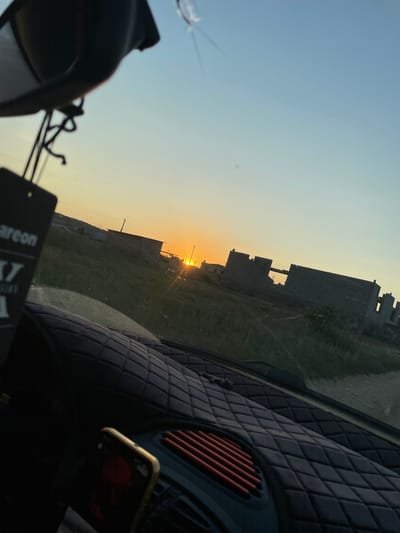 The media file depicts a view from inside a vehicle looking outwards during what appears to be sunset. The scene is set in a semi-urban or rural area, with unfinished brick buildings suggesting construction or a developing neighborhood. The sky is a gradient of pale blue to a warm orange near the horizon, indicative of the golden hour. Green grass and some vegetation are visible in the foreground and around the buildings.
Inside the vehicle, the dashboard is partially visible, featuring a diamond-patterned texture and an air vent with red slats. A smartphone is mounted on the dashboard, displaying a dark screen with some faint elements, possibly notifications or a background image. In the lower left corner of the frame, a dark object, possibly a car accessory or a hanging air freshener, is partially visible. A small tag with visible text hangs from this object.
The overall mood is serene and contemplative, possibly reflecting a moment of travel or observation during the end of the day. The sun is low on the horizon, casting a warm glow and long shadows. The image composition suggests a personal perspective, likely taken by the driver or a passenger. The location context provided, Buynaksk, Russia, implies the setting is within this geographical area.
Visible text on the tag in the lower foreground includes "areon" and a stylized "X7".