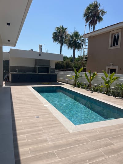 This outdoor scene captures a modern residential property in Moni Community, Cyprus, featuring a swimming pool and an outdoor kitchen area. The swimming pool, rectangular in shape, is filled with turquoise water and surrounded by light-colored tiled decking. To the left of the pool, a covered outdoor kitchen is visible, equipped with dark-colored cabinetry, a countertop, and a built-in barbecue grill with a stainless-steel chimney. The background includes a two-story building with a beige facade and a tiled roof, flanked by several tall palm trees. A white horizontal fence separates the property from neighboring areas. The sky is clear and blue, indicating fair weather. The overall impression is one of a newly constructed or renovated outdoor living space designed for leisure.