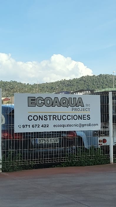 An outdoor scene captures a white banner sign for "ECOAQUA .tec PROJECT CONSTRUCCIONES" in the Polígon Industrial De Calvià, Spain. The sign prominently displays the phone number "971 672 422" and email "ecoaquatecnic@gmail.com". The banner is affixed to a dark-colored metal grid fence. Behind the fence, the rear of a dark-colored vehicle is visible, featuring a license plate that reads "B 756 KSM". Green foliage grows along the base of the fence, separating it from a dark gray paved surface in the foreground. In the mid-ground and background, multiple distant green, tree-covered hills rise under a bright blue sky dotted with scattered white cumulus clouds. No individuals are present in the image.