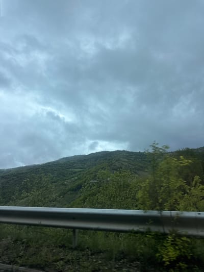 The image shows a landscape taken from a moving vehicle, likely a car, as evidenced by the guardrail on the right side of the frame. The left side of the image is dominated by a cloudy, overcast sky. To the right, a mountain covered in dense green vegetation rises sharply. The location is potentially near Liaskovo, Bulgaria, based on the request, although specific landmarks to confirm this are absent. No people or distinct subjects besides the natural environment are identifiable. The action is implied movement of the vehicle taking the photo.
