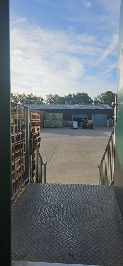 The image shows a view from inside a building in Gravesham, United Kingdom, possibly a loading dock. Outside, there's a concrete yard with stacked wooden pallets and crates. A warehouse-style building is visible in the background with closed roller doors. There are a few pieces of equipment near the building, possibly a small construction vehicle. The sky is blue with some clouds. Part of a metal ramp or loading platform is visible in the foreground with a diamond plate surface.