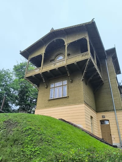 The image shows a multi-story wooden house with green siding and a yellow base, located on a grassy incline. The house features a prominent balcony with ornate railings and decorative eaves. A window with multiple panes is visible on the lower level, and an arched window is present on the upper level. To the left of the house, there are trees and a lamppost. At the base of the grassy incline, there appears to be a historical cannon. The sky is overcast, suggesting it might be raining or foggy, with visible water droplets in the air. This property is known as the Rendas Manor in Kuldīga, Latvia.