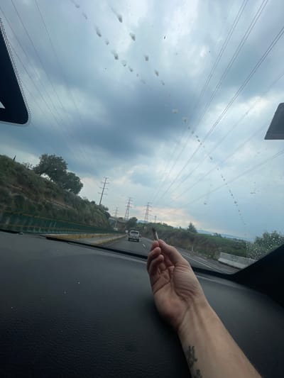 The image, taken from inside a vehicle, shows a person's hand holding a rolled cigarette. The hand is positioned in the foreground, with a tattoo visible on the forearm. The view through the windshield reveals a cloudy sky with numerous power lines crisscrossing the frame. Outside the vehicle, a highway is visible with traffic, including a white truck. The roadside is lined with greenery and rocky hills, with power towers dotting the landscape. The overall scene suggests a journey on a highway under a cloudy sky, with the presence of a lit cigarette indicating a moment of pause or relaxation during transit. The location is identified as Ciudad López Mateos, Mexico, based on the context provided.