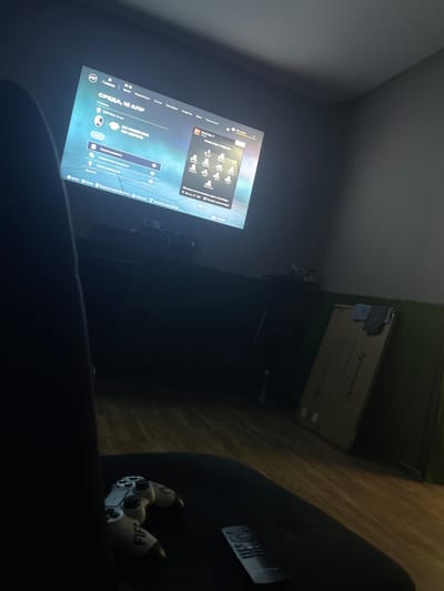 A dimly lit room features a large television screen displaying a soccer video game interface, likely FIFA. The screen shows team information, player avatars, and menu options in Russian. A black gaming chair is partially visible in the foreground, with a white and gray video game controller resting on its seat. A silver remote control is also present on the seat. In the background, a dark cabinet or entertainment unit is visible, with various electronic devices and cables. To the right of the screen, a large cardboard box with the "BRAUBERG" logo is leaning against a wall, and a piece of green artificial turf is visible near the floor. The floor appears to be made of wooden planks. The scene is consistent with a home entertainment setup, possibly in Nevinnomyssk, Russia, given the Russian language interface on the screen.