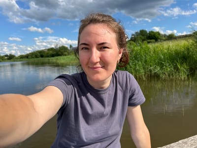 This is an upside-down, eye-level selfie of a young woman taken outdoors. She is wearing a dark purple t-shirt. Her face is the central focus, and she has light brown hair, blue eyes, and fair skin. She appears to be smiling slightly, with a relaxed and pleasant expression. Her left arm is visible, bent and extended towards the camera.
The background shows a body of water reflecting the sky and a line of green reeds or tall grass along the bank. Beyond the reeds, there are trees and a bright blue sky with fluffy white clouds, suggesting it is daytime with good weather. The overall scene evokes a peaceful, natural setting, likely near a lake or river. There is no visible text in the image. The orientation suggests the photo was taken while lying down or in an inverted position.