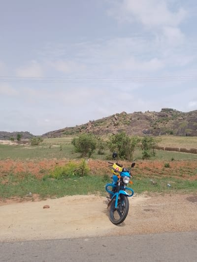 A medium shot shows a blue motorcycle parked on a dirt road in a rural setting. Behind the motorcycle, the land stretches out to a hill covered in rocks and sparse vegetation. In the background, the sky is overcast and power lines are visible.
There is no indication that this image is related to Barakin Tenti, Nigeria, unless further information is provided.