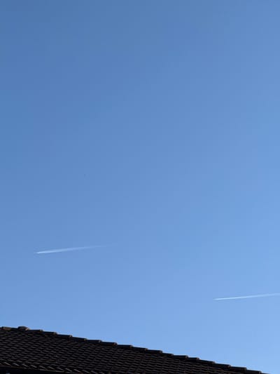 The image shows a clear blue sky with two visible contrails from airplanes. The edge of a dark tiled roof occupies the right side of the frame. The location is Zhaltusha, Bulgaria. No people are visible.