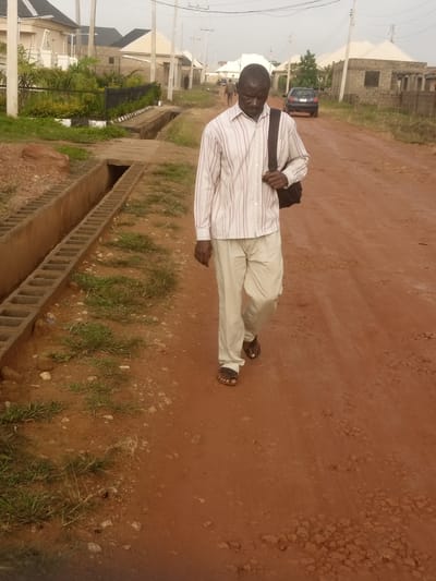 An adult male wearing a light-colored striped long-sleeved shirt and light-colored pants walks on a dirt road in what appears to be a developing urban area. He is carrying a dark-colored bag over his left shoulder. The man is wearing sandals. A concrete drainage ditch runs alongside the road to the left of the frame. In the background, there are houses, some appearing to be under construction, and utility poles. A dark-colored car is visible further down the road. The ground is a reddish-brown dirt, with patches of sparse green grass and scattered small rocks. The sky is overcast. The scene suggests an outdoor environment in a town or city, possibly Russo, Nigeria, given the characteristic red earth and architectural style of the buildings.