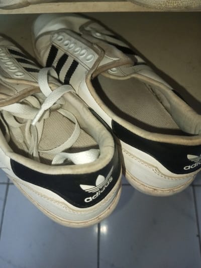 Two well-worn Adidas sneakers sit side-by-side. The shoes are predominantly white with black stripes and heel trim, featuring the Adidas logo prominently on the heel. The laces are loosely tied on the left shoe, while the right shoe is unlaced. Both sneakers exhibit signs of wear, with dirt and scuff marks visible on the soles and uppers. They rest upon light gray tiles.