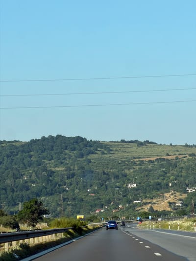 An eye-level, vertical, medium shot captures a highway scene under a clear, light blue sky with the distant, rolling hills of Eleshnitsa, Bulgaria. In the foreground, a two-lane asphalt highway stretches forward, bordered by a guardrail on the left and a grassy shoulder with reflective markers on the right. Several vehicles are visible on the highway, including a blue car in the left lane and a yellow truck in the right lane further ahead. The middle ground features a dense, green hillside dotted with scattered houses and power lines, indicating a semi-rural or suburban setting. The background consists of the vast, unbroken expanse of the sky. The overall lighting suggests daytime with bright sunlight.