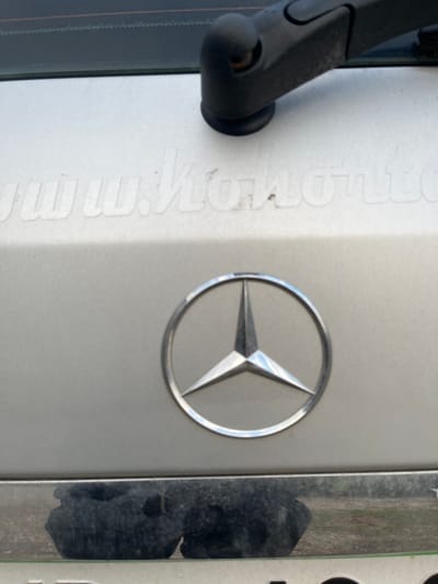 The image displays a close-up, angled view of the rear section of a light-colored vehicle, specifically a tailgate or trunk lid, located in Brežice, Slovenia. A chrome Mercedes-Benz three-pointed star emblem is centrally positioned in the lower half of the frame. Above the emblem, faded white block lettering is partially visible on the vehicle surface, beginning with "www.Ko" and continuing with indistinct characters that are obscured by dust or wear. In the upper right quadrant, a black rear windshield wiper arm is present, positioned below a dark-tinted rear window. Below the Mercedes emblem, a horizontal chrome trim strip reflects a dark, indistinct outdoor ground area, suggesting either vegetation or dirt. The vehicle's surface shows a thin layer of dust and grime. No individuals or explicit actions are depicted.