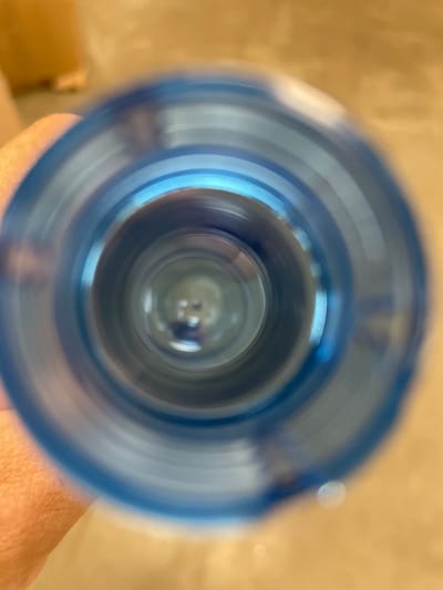 The image shows a close-up, distorted view looking down into the opening of a blue plastic bottle. The perspective is from the viewpoint of someone holding the bottle, with a portion of their hand visible on the left side of the frame, out of focus. The background, visible around the bottle opening and through it, is a blurred, warm-toned surface, possibly a wooden floor or wall. Concentric rings of blue plastic are visible as the sides of the bottle's opening. The center of the opening is a dark, blurry circular area, with faint reflections suggesting a shiny inner surface or liquid. The scene does not contain any identifiable landmarks or elements that would specifically place it in Терволово, Russia.