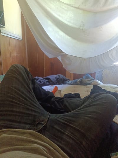 The scene is an interior setting, presumably a bedroom. The primary subject is a person lying on a bed, viewed from a first-person perspective, focusing on the legs clad in dark-colored jeans. One foot, clad in an orange sock, is visible.
The bed has multiple layers of bedding including a quilt. Above, a white sheet is draped, forming a makeshift canopy. The background includes a wood-paneled wall, a window with light filtering through, and what appears to be a room fan.
The location is assumed to be Park Hill, United States, but there are no identifiable landmarks or details in the image to confirm this. No specific actions or interactions are evident other than the person lying on the bed.