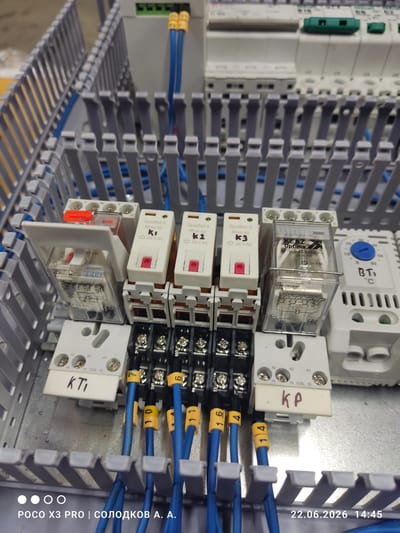 This is a close-up, high-angle photograph of an electrical control panel. The panel is filled with various components, including relays, terminal blocks, and wiring. The relays are predominantly white and clear plastic, with some featuring red push-buttons or levers. Blue wires with yellow wire markers are neatly organized and routed through grey plastic channels.
One relay is labeled "KEAZ Optima OptiRel 6 RP55-34" and shows electrical specifications like "240 VAC" and "6A 30V DC". Another relay has "K1", "K2", and "K3" printed on its casing, along with "24 V AC". A component with a blue dial and markings "BT" and "°C" suggests a temperature control or sensor.
Several relays have handwritten labels: "KP" and "KTI". The wires are marked with yellow tags showing numbers such as "16", "4", "1", and "0". The overall setting appears to be an industrial or technical environment.
The bottom right corner of the image contains overlaid text, likely from a device's camera, indicating "POCO X3 PRO | СОЛОДКОВ А. А." and the date "22.06.2026 14:45". There are also three white circles below this text, possibly status indicators. The location context is Uryupinsk, Russia. The image is well-lit, suggesting an indoor setting, and there is no indication of the time of day or weather conditions. The focus is entirely on the technical components within the panel.