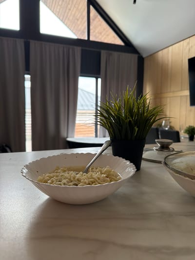 A close-up, eye-level shot captures a bowl of ramen noodles with a fork resting in it, placed on a marble-patterned countertop. To the right, a potted green plant adds a touch of life to the scene. In the background, a modern interior features large triangular windows with grey curtains, a wooden-paneled wall, and other dining elements like glasses and a lid from another pot. The overall impression is one of a casual meal in a contemporary living space. The location is identified as Semiluki, Russia.