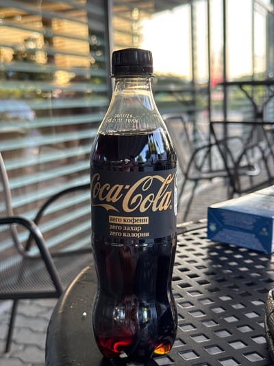 The image depicts a close-up, eye-level shot of a plastic bottle of Coca-Cola Zero Sugar, standing upright on a dark, patterned outdoor table. The bottle is mostly filled with a dark liquid and has a black cap. The label is primarily black with gold lettering, displaying the Coca-Cola logo and text in Cyrillic. The text appears to read "zero кофеин" (zero caffeine), "zero захар" (zero sugar), and "zero калории" (zero calories).
The background is blurred, suggesting a shallow depth of field. Through the bottle, reflections of outdoor elements are visible, including horizontal blinds and greenery, indicative of an outdoor seating area. To the right of the bottle, a partially visible blue box with some graphics is also on the table. In the background, outdoor seating furniture, possibly chairs and tables, are faintly discernible. The overall lighting suggests it is daytime, with sunlight filtering through the background elements, creating a warm, slightly hazy atmosphere. There are no people visible in the image. The scene is set in an outdoor seating area, potentially a cafe or restaurant, in the city of Razliv, Bulgaria.