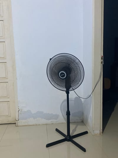 The image shows a black standing fan on a white wall. The fan has a round head with a black grate and a white logo in the center. The fan is on a black pole with a tripod base. The fan is plugged into a white power strip that is attached to the top of the wall. The wall is painted white and has some water stains on it. There is also a dark blue object in the background. The image appears to be taken indoors, possibly in a bedroom or living room. The lighting is dim, suggesting it may be nighttime or the room is not well-lit. The overall impression is of a simple domestic setting. There is no visible text in the image.