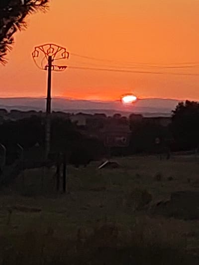 An outdoor, medium-shot, blurry photograph depicts a sunset with a bright orange sky. In the foreground, a dark, silhouette of a power pole with insulators and wires is visible on the left side of the frame. The horizon line is marked by a distant, dark silhouette of hills and trees. The sun, a bright white circle with a reddish glow, is setting just above the horizon. The lower portion of the image is dark and blurry, showing a field with some indistinct objects and structures. The image appears to have been taken in or near the city of Sobradillo, Spain.