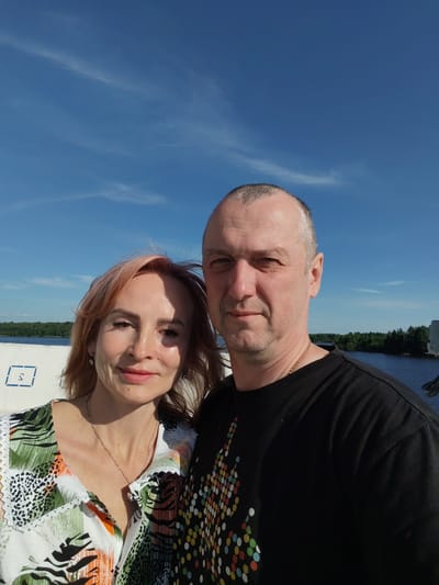 A man and a woman are posing for a selfie with a bright blue sky and wispy clouds in the background. The woman is on the left, with shoulder-length, reddish-blonde hair, and is wearing a white shirt with a colorful abstract print and a delicate necklace. She has a slight smile and appears to be looking at the camera. The man is on the right, with short, graying hair and a light beard. He is wearing a black t-shirt with a colorful circular pattern on the front and a gold chain. He is looking directly at the camera with a neutral expression.
In the background, a body of water, possibly a river, is visible with lush green trees lining the opposite bank. To the left of the couple, there is a white structure with a blue marking that resembles the letter "S" enclosed in a square. The lighting suggests it is daytime, likely midday, given the bright sunlight and clear sky. The overall mood of the image is casual and relaxed, as if the couple is enjoying an outdoor activity.
The location context provided is Nova, Russia, which could be a clue to the setting, possibly a waterfront area or a vessel on the water. No specific event is discernable beyond the selfie being taken.