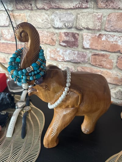 An ornate, carved wooden elephant figurine is displayed on a black surface against a brick-patterned wallpaper. The elephant is adorned with two bracelets: a multi-strand turquoise beaded bracelet is wrapped around its trunk, and a single strand of white pearls encircles its body. To the left of the elephant, several watches are visible on a decorative gold leaf-shaped placemat, including one with a white band and another with a dark blue band. A black clutch bag and a red box are also present on the surface. The lighting suggests an indoor setting. The city of Рубцово, Russia, is not visually represented in this image.