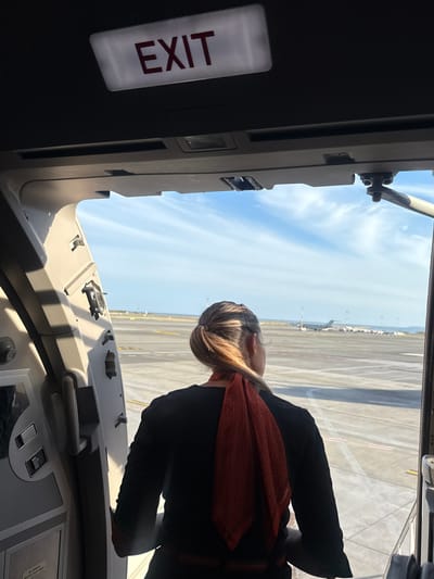 The image provides an interior view from an easyJet aircraft, looking outwards through an open door onto the tarmac of Nice Côte d'Azur Airport (NCE) in Saint-Laurent-du-Var, France. An illuminated "EXIT" sign with red lettering is affixed to the dark ceiling of the aircraft, positioned above the open doorway. The light-colored interior frame of the aircraft door is visible, showing various latches and structural elements. In the immediate foreground, with her back to the viewer, stands a person wearing a black long-sleeved top and a textured orange-red scarf, which bears the "easyJet" logo on its lower right portion. Her light brown hair is styled in a ponytail, and she is observing the exterior airport environment. Beyond the aircraft door, a expansive concrete tarmac with white and yellow markings extends into the distance. Several airplanes, including a distinct dark-colored business jet, are visible parked or positioned on the tarmac in the mid-ground. Various airport poles and infrastructure are scattered across the field. In the far background, a clear blue sky with scattered wispy clouds meets the horizon, where the Mediterranean Sea is discernible. The scene is brightly illuminated by daylight.