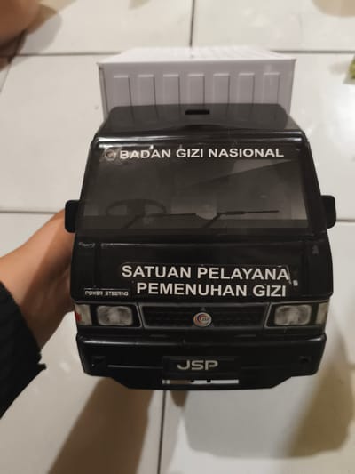 A black toy pickup truck with a white box body is held from its left side by a person's left hand. The truck faces the viewer, positioned on a light-colored, tiled floor. The windshield displays a reflection, and a steering wheel and dashboard details are visible through it. White text on the upper part of the cabin reads "BADAN GIZI NASIONAL," which translates to "NATIONAL NUTRITION AGENCY." Below the windshield, on the lower front of the cabin, more white text states "SATUAN PELAYANA PEMENUHAN GIZI," translating to "NUTRITION FULFILLMENT SERVICE UNIT" (with a slight grammatical error in the original Indonesian). A small rectangular plaque near the left headlight features the words "POWER STEERING." The truck's grill prominently displays a circular "JSP" logo, and the letters "JSP" are also present in white text on the front bumper. The ambient lighting casts soft shadows of the truck and hand onto the floor, indicating an indoor setting.