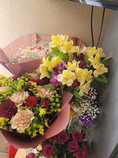 The image displays a vibrant collection of several flower bouquets, arranged together, likely on a counter or table within an indoor setting in Šilutė, Lithuania.
The most prominent bouquet, positioned centrally and to the left, features bright yellow and deep purple alstroemeria wrapped in pink paper. To its right, another large bouquet showcases a mix of red and light pink/peach carnations, deep red chrysanthemums or similar flowers, bright yellow solidago (goldenrod), and small green berries, all wrapped in a maroon paper.
Towards the top right, a bouquet with white baby's breath (gypsophila), purple freesias, and a few yellow flowers is visible, encased in clear plastic wrapping. Below and to the right of this, a bouquet of deep red roses with green foliage can be seen, also partially wrapped in maroon paper. Another bouquet, partially visible on the far left, includes light pink and white roses along with baby's breath, wrapped in pink paper.
The background consists of a light brown or beige surface with a subtle, swirling patterned wallpaper. A dark, reflective metallic edge, possibly part of a refrigerator or cabinet, is visible along the top left side of the image. A small red object is partially visible at the bottom right. No activity or people are shown, and there is no visible text. The image captures the bouquets from a slightly elevated angle, highlighting their diverse colors and textures.
