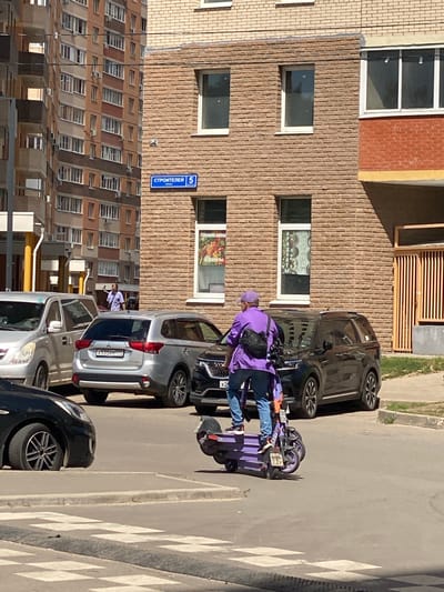 In Kotelniki, Russia, under the bright daylight and clear weather, a person in a vibrant purple jacket and matching hat is on an electric scooter. The scooter itself is also a striking purple, parked on a paved street marked with a pedestrian crossing. The scene is set against a backdrop of a multi-story building with a facade of light brown brick, featuring numerous windows, some with white frames, and others with darker glazing. To the left, another building with a similar brick pattern is visible. Several cars are parked along the street; a dark-colored car is in the foreground, and a silver SUV is parked behind it. A blue street sign with white text, reading "СТРОИТЕЛЕЙ 5," indicating the street name and building number, is affixed to the brick building. The overall impression is of an everyday urban scene in a residential area.