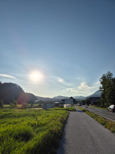 The image depicts a scenic view in Niederndorf, Austria, on a clear day with a bright sun. The foreground features a lush, green grassy field and a paved pathway. To the right, a road runs parallel to the pathway, with a few cars visible. Beyond the road, there are houses and trees, and in the background, rolling hills and mountains are silhouetted against the sky. The sun is low in the sky, casting a warm glow and lens flare across the scene. The overall impression is one of a peaceful rural landscape.