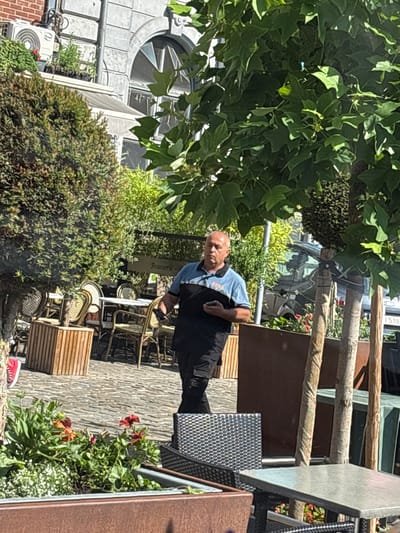 A man in a blue shirt and black trousers stands in front of a brasserie. He is holding a smartphone in his right hand and appears to be looking at it. The brasserie has outdoor seating with wicker chairs and tables. There are also planters with red and orange flowers. A large tree with green leaves shades part of the scene. In the background, there is a building with a circular window and an air conditioning unit. The scene appears to be set during the daytime, likely on a sunny day given the shadows. The overall impression is that of a casual outdoor dining setting.
