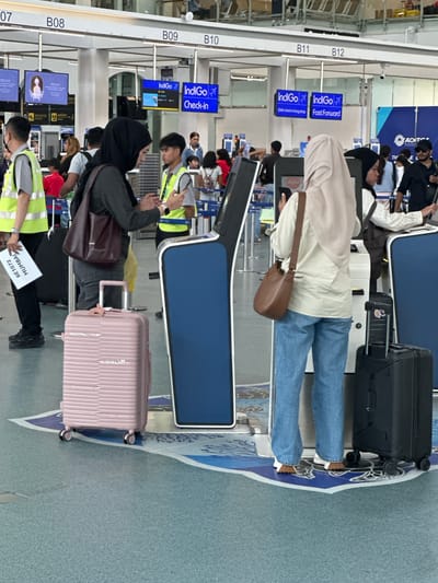 The image depicts an indoor scene at an airport in Tambon Mai Khao, Thailand, likely a check-in area. Several individuals are present, appearing to be passengers and airport staff. The foreground features two large suitcases, one pink and one black, positioned on a patterned floor. In the mid-ground, two women wearing hijabs are interacting with self-service check-in kiosks. One woman, on the left, is looking down at her phone, while the other, on the right, is engaged with the kiosk. Behind them, airport staff in high-visibility vests can be seen directing passengers and managing the check-in process. Overhead signage indicates various check-in counters labeled with numbers and airline names, such as "IndiGo." The environment appears well-lit and functional, consistent with an airport terminal.