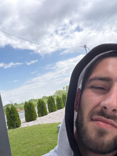 A close-up selfie captures a man with a beard, wearing a hooded sweatshirt. The background reveals a landscape with a partially cloudy sky, green grass, and a line of bushes or shrubs. A wire and a pole are visible against the sky. The location appears to be Yoakim Gruevo, Bulgaria, based on the likely origin of the photograph.