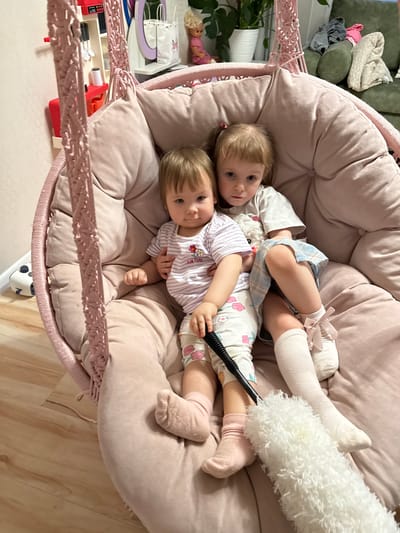 Two young children are lying together in a large, round, cushioned, light pink hanging chair. One child is wearing white knee-high socks with ribbons, a light-colored dress with blue plaid, and a light-colored shirt. The other child is wearing pink socks and white pajamas with pink flowers, paired with a striped shirt. This child is holding a black-handled duster. The location in Krapivka, Russia, is not apparent from the image. The chair is hanging indoors with a white wall and a light wood floor. In the background is a green couch and various toys.