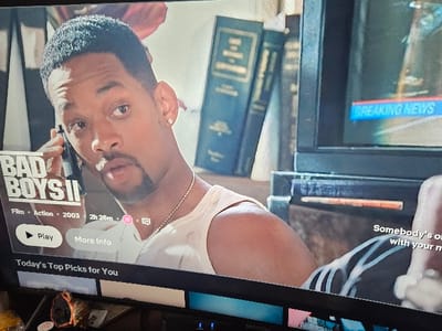 The image displays a television screen presenting a streaming service interface, showing a still from the film "Bad Boys II." The central figure on the screen is actor Will Smith, a Black male with short dark hair and a goatee, wearing a white tank top and a thin silver chain necklace, with a gold stud earring in his left ear. He is depicted holding a black telephone-like object to his left ear, looking towards the right with his mouth slightly open. In the background of the movie scene, dark bound books are visible on a shelf to the right, adjacent to a smaller television displaying "BREAKING NEWS" in blue text.

Overlays on the screen identify the content as "BAD BOYS II," a "Film - Action - 2003" with a duration of "2h 26m." A pink circular icon and another small white icon are present next to the duration. Below the title, "Play" and "More Info" buttons are displayed. White subtitles on the right side of the screen partially read, "Somebody's o[ut] with your m[oney]". A dark bar near the bottom of the screen reads "Today's Top Picks for You," above a row of partially visible content thumbnails.

The physical black frame of the television is visible at the bottom and partially on the right. Blue indicator lights are discernible on the bottom edge of the television's frame. Beneath the television, a dark, textured surface supports the device. On this surface, to the left, an irregularly shaped orange-brown object and some translucent white material are present.

The image itself contains no discernible information regarding its specific location within the Cherwell District, United Kingdom.