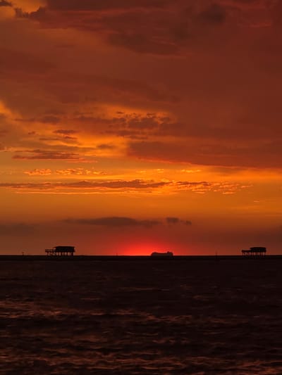 This image depicts a dramatic sunset over the Adriatic Sea, likely viewed from Chioggia, Italy, a coastal city known for its picturesque lagoons and fishing traditions. The sky is ablaze with vibrant hues of orange and red, with a sliver of the sun's disc peeking through the horizon, casting a fiery glow. Wispy clouds, illuminated from below, add texture and depth to the celestial canvas.
In the foreground, the dark, choppy surface of the sea stretches across the frame, reflecting the somber tones of the sky. A thin, dark line on the horizon separates the sea from the sky, and on this line, two silhouetted structures are visible. The structure on the left is a stilted building, possibly a fishing hut or a small dwelling, raised on wooden posts above the water. To its right, a flatter, more elongated silhouette suggests a pier or a breakwater, with a solitary post marking its presence. Further to the right, a second, similar stilted building is also visible, situated on the distant horizon. The overall impression is one of serene isolation and the raw beauty of nature.