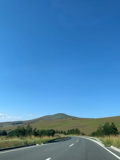 The image depicts a clear blue sky over a winding asphalt road in Cajetina Municipality, Serbia. The road curves to the left, with a metal guardrail lining its right side. Beyond the road, a field of tall, dry grass and scattered evergreen trees are visible. Further in the background, rolling green hills rise, some with patches of darker vegetation and what appear to be distant buildings or settlements. The overall scene conveys a sense of open, rural landscape under a bright, sunny day. There are no people or animals visible in the image.