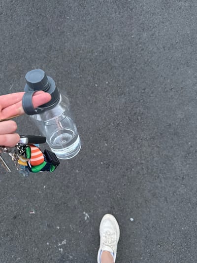 A person is walking on asphalt. In the foreground, a hand holds a clear water bottle with a gray lid and a gray loop. Attached to the bottle's loop is a keychain with several keys and colorful charms. The person is wearing a white sneaker. The background is an out-of-focus view of asphalt. The setting appears to be an outdoor public space.