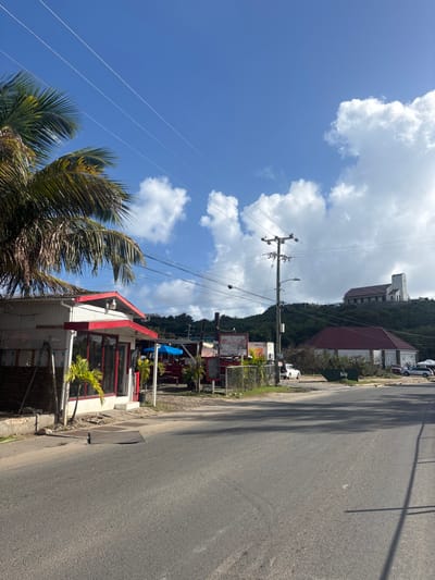 The image depicts a roadside scene in Spanish Town, British Virgin Islands, under a clear blue sky with scattered white cumulus clouds.
In the foreground, a paved asphalt road extends horizontally across the frame. To the left, a large palm tree with green and brownish fronds partially obscures the upper left corner. Below it, a single-story commercial building with a white facade and a red gabled roof is visible. This building features large glass panel windows and red trim, with two small green palm saplings positioned in front.
Moving right along the roadside, an outdoor seating area with a blue umbrella and red furniture is partially visible, adjacent to a sign that reads "CHE MIMI'S KITCHEN." This area is fenced off by a green chain-link fence. Behind the fence, a white utility pole stands upright, supporting multiple power lines that traverse the sky. A white compact SUV is parked near the base of the utility pole, off the main road.
In the midground, a large building with a dark red or brown gabled roof and white walls is situated on a lower elevation, to the right of the commercial area. The background features a prominent, green, vegetated hill. Perched atop this hill, a large, light-colored building with a distinctive architectural design, including a tall, pointed section, is clearly visible against the sky. The scene is illuminated by direct sunlight, casting shadows. No individuals are visible.