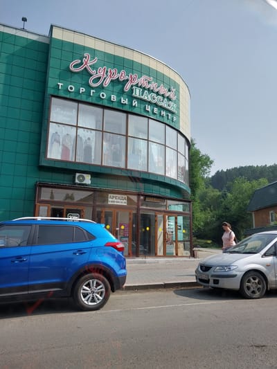 A wide, low-angle exterior shot captures the "Курортный Пассаж" shopping center on a sunny day in Belokurikha, Russia. The building features a curved facade with teal-colored tiled sections on the upper floors and a large, multi-pane window showcasing mannequins dressed in clothing. The prominent red and white neon sign "Курортный Пассаж" curves across the top of the building, with smaller text below reading "ТОРГОВЫЙ ЦЕНТР".

At the street level, glass doors lead into the shopping center. One door has a sign advertising "АРЕНДА" (Rent) with a phone number. Two cars are parked on the street in front of the building: a bright blue SUV on the left and a silver sedan on the right. A woman with brown hair, wearing a light pink shirt and blue jeans, is walking away from the shopping center towards the silver car. The environment is a typical street scene with a sidewalk, road, and a backdrop of lush green trees and hills.