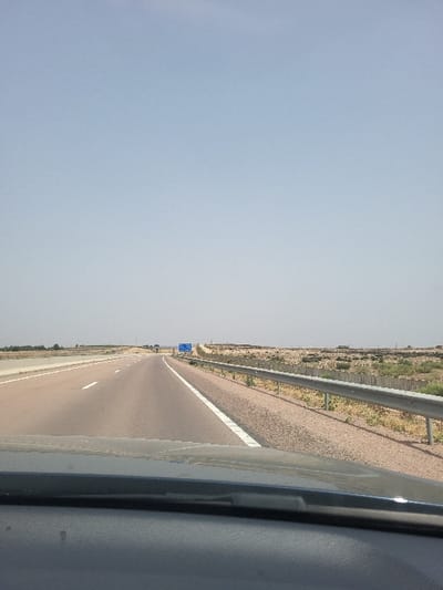 The image displays a forward-facing view from within a vehicle onto a multi-lane asphalt highway in Oulad Ghanem, Morocco. The road features at least two visible lanes marked by white dashed lines, with a solid white line bordering a wide gravel shoulder on the right. A silver metal guardrail runs parallel to the shoulder. The environment consists of flat, arid, reddish-brown terrain with sparse, low-lying dry vegetation under a clear, pale blue sky. In the far distance, a blue overhead road sign is visible spanning the highway, with what appears to be an overpass or bridge structure beyond it. No individuals or other active subjects are present in the frame.