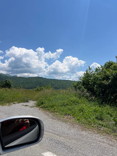 This is a first-person perspective photograph taken from within a vehicle, showing a scenic mountain landscape under a bright blue sky with scattered white cumulus clouds. In the foreground, a gravel road is visible, with green grass and bushes lining its side. The driver's side mirror reflects the interior of the car, including the arm of a person wearing a red shirt and holding a phone. The background features rolling green hills covered in dense forest, with some parts of the hills fading into a misty haze. The overall impression is one of a clear, sunny day in a rural, mountainous region. The location is identified as Bakovići, Montenegro.
