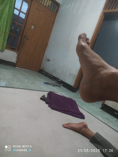 The image shows the interior of a home in Kebumen, Indonesia, with a white tiled floor. A person is lying on a rug, with their right foot raised towards the ceiling, and the bottom of their right leg is in view. There is a wooden door to the left of the image, and a wall to the right. A purple towel lies on the rug, and a shadow of a small object can be seen near it. The timestamp on the bottom right corner of the image indicates the date and time as 21/05/2025 17:36.