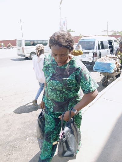 This image captures a daytime street scene in Communauté rurale de Payar, Senegal. In the foreground, a woman with dark, short curly hair is walking on a paved surface. She wears a short-sleeved, patterned green and black top with matching trousers, a black wristwatch on her left wrist, and carries a black leather handbag in her right hand and a black plastic bag in her left hand. Her gaze is directed downwards.
Behind her, to the left, another person is partially visible from behind, wearing a light-colored hooded top and light-colored trousers, also walking. Further right in the midground, a white utility van or minibus with a damaged rear bumper is stopped or parked; its rear door is open, revealing cargo. Adjacent to the van, a two-wheeled pushcart is laden with goods, including a pile of nuts or similar items and a blue tarp covering other contents. A person is partially visible near the cart.
In the background, a wide, paved road shows vehicular traffic, including a white minibus moving from left to right and a person on a bicycle or moped. The sky is clear and bright, indicating strong daylight. Utility poles and distant structures are visible on the horizon.