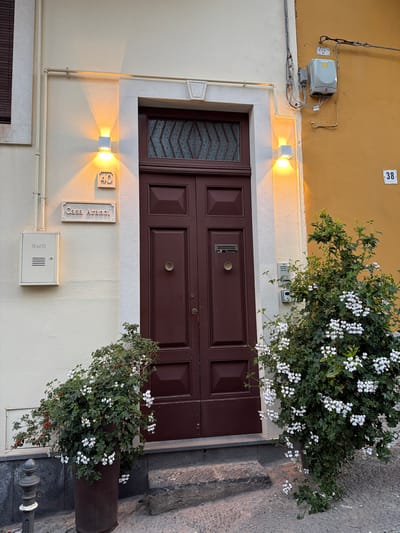 The image depicts the entrance to "Casa Aranci" located at number 40, in Acireale, Italy. The entrance features a brown, double-paneled door with a small window at the top. Two outdoor lamps flank either side of the door illuminating it with a warm glow. A plant with white flowers partially obscures the upper right portion of the door. A gas meter is mounted on the wall below the house name, while another utility box is located above the door on the yellow wall of the adjacent building, marked with the number 38. A narrow section of sidewalk is visible next to the entrance.