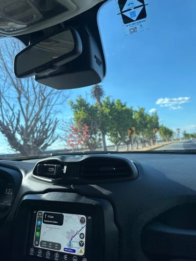 The view is from inside a car, looking through the windshield. A rear-view mirror is positioned in the top left. The dashboard is visible with an air vent and a screen displaying a map. The navigation shows that Caseta de Cobro is 11 km away. Trees are visible outside. The sky is blue with some clouds. The display screen shows icons for various applications like radio and media. The number 122 is visible on the dashboard.