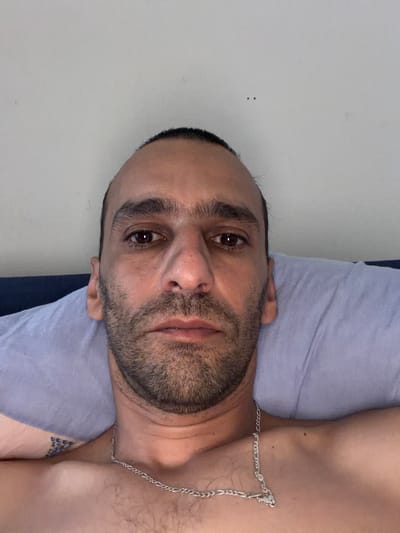 The image is a close-up, overhead shot of a man lying on his back on a pillow, possibly in a bedroom. The man has dark hair, dark eyebrows, and a beard. He is wearing a silver chain around his neck. His eyes are open and looking directly at the camera. The background consists of a light-colored wall. The overall impression is that of a casual, indoor portrait. There is no information available about the specific location within Schaerbeek - Schaarbeek, Belgium.