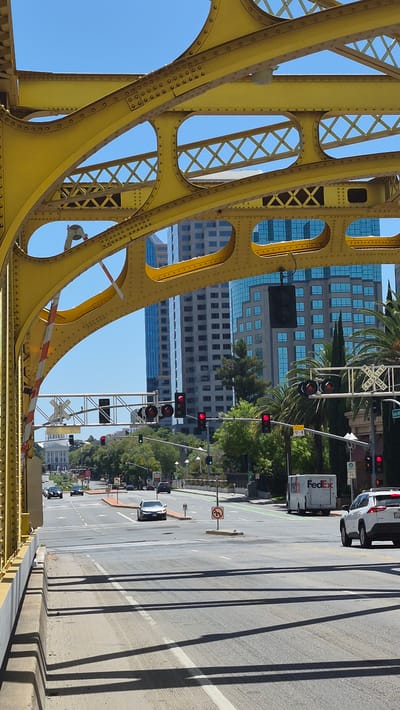 A view from a yellow truss bridge over a street in West Sacramento, United States of America, shows traffic on a multi-lane road beneath the bridge. Modern skyscrapers and trees are visible in the background. Several cars are on the road, including a white SUV and a silver sedan. A white FedEx truck is also present. Traffic lights are visible, with some showing red and others green. A railroad crossing sign is also present. The sky is clear and blue. The bridge's structural elements, painted yellow, dominate the foreground.